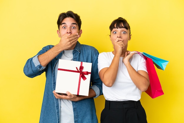 Junges Paar mit Einkaufstüten und Geschenk isoliert auf gelbem Hintergrund, der den Mund mit den Händen bedeckt, weil es etwas Unangemessenes gesagt hat