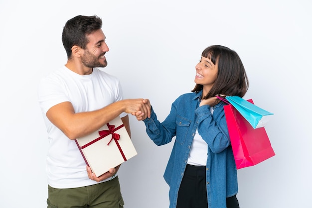 Junges Paar mit Einkaufstüten und Geschenk isoliert auf weißem Hintergrund Händeschütteln nach einem guten Geschäft