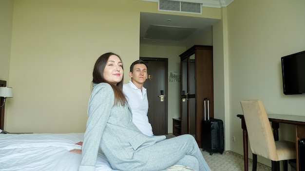 Junges Paar mit Koffer betritt das Hotelzimmer und setzt sich aufs Bett