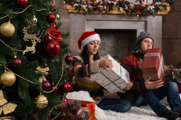 Junges Paar mit lustigen Hüten und Pullovern sitzt auf einem Teppich vor dem geschmückten Kamin und dem Weihnachtsbaum, der seine Weihnachtsgeschenke auspackt