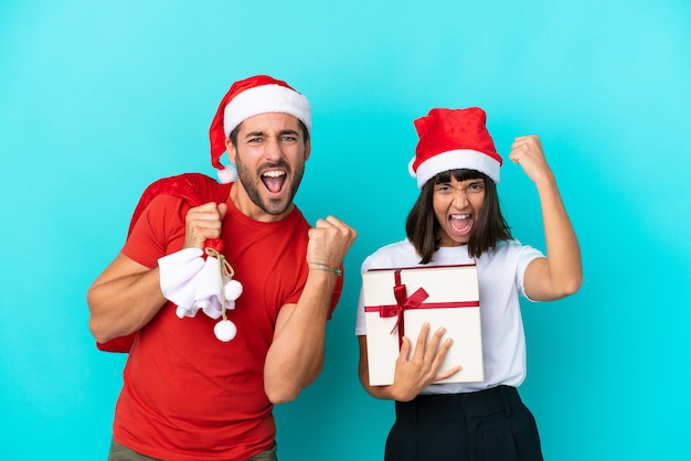 Junges Paar mit Weihnachtsmütze verteilt Geschenke isoliert auf blauem Hintergrund und feiert einen Sieg