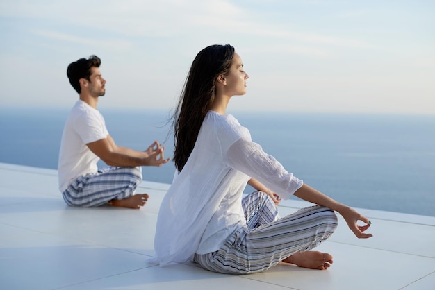 junges Paar praktiziert Yoga bei Sonnenuntergang in der modernen Hausterrasse mit Meer und Sonnenuntergang im Hintergrund