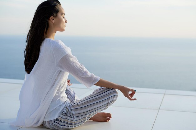 junges Paar praktiziert Yoga bei Sonnenuntergang in der modernen Hausterrasse mit Meer und Sonnenuntergang im Hintergrund