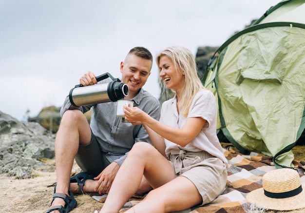 Junges Paar Reisende in den lässigen Outfits mit Zelt Mann gießt Tee aus Thermoskanne Wochenendausflug zum lokalen Tourismus