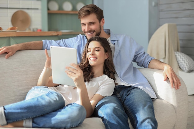 Junges Paar sieht sich Medieninhalte online auf einem Tablet an, das auf einem Sofa im Wohnzimmer sitzt.