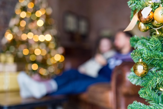 Junges Paar sitzt auf dem Sofa in der Nähe des geschmückten Weihnachtsbaums im gemütlichen Loft verschwommenes Bild