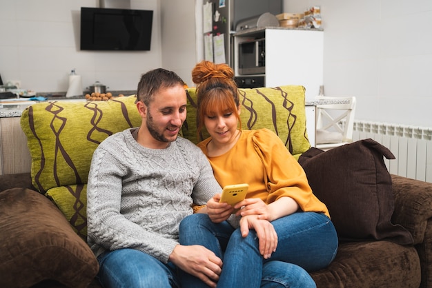 Junges Paar sitzt zu Hause auf der Couch und schaut lächelnd und glücklich auf ein Smartphone zusammen mit der Küche im Hintergrund