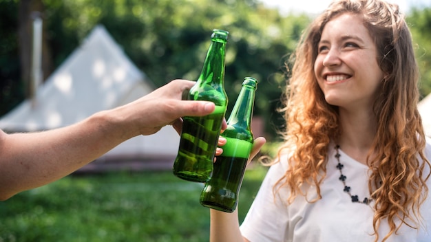 Junges Paar trinkt Bier, lächelt, klirrende Flaschen. Grün herum. Glamping