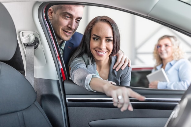 Junges Paar überprüft die Eigenschaften des neuen Autoinnenraums, während der Verkaufsleiter im Autohaus beiseite steht
