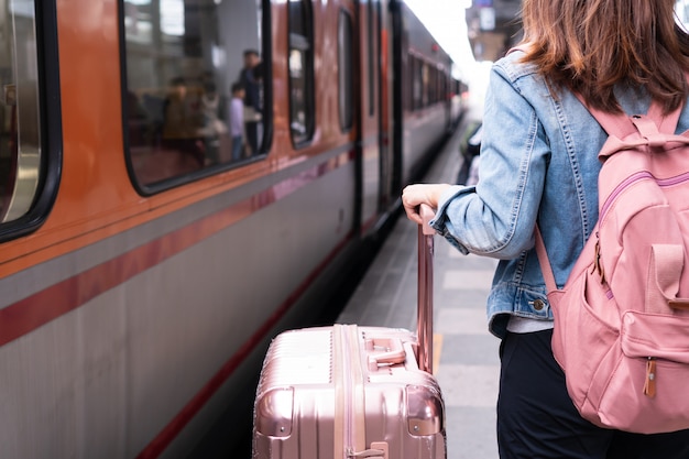 Junges reisendes Mädchen in der Jeansjacke mit der rosa Tasche und dem Gepäck, die auf den Zug auf dem Bahnsteig, Kopienraum, Reise- oder Transportkonzept warten