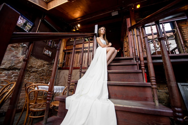 Foto junges romantisches elegantes mädchen im langen weißen fließenden kleid, das auf der treppe innen aufwirft