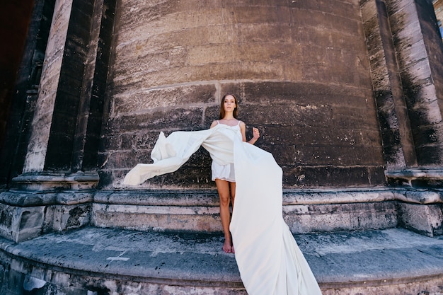 Junges romantisches elegantes Mädchen im langen weißen fließenden Kleid, das über alte Steinmauer aufwirft