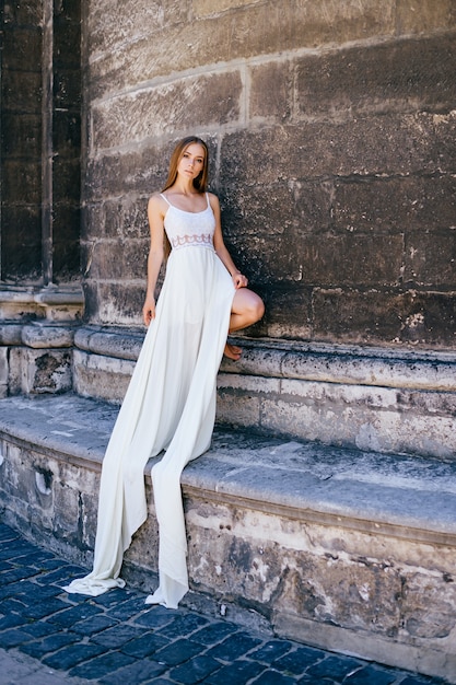 Junges romantisches elegantes Mädchen im langen weißen Kleid, das über alte Steinmauer aufwirft