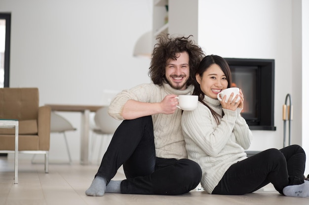 Junges romantisches multiethnisches Paar sitzt zu Hause auf dem Boden vor dem Kamin, redet und trinkt Kaffee an kalten Wintertagen