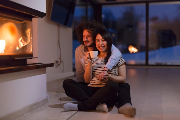 Junges romantisches multiethnisches Paar sitzt zu Hause auf dem Boden vor dem Kamin, redet und trinkt Tee in kalter Winternacht