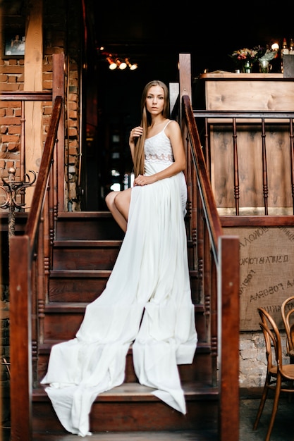 Junges romantisches nachdenkliches Mädchen im langen weißen Kleid, das auf der Treppe sitzt