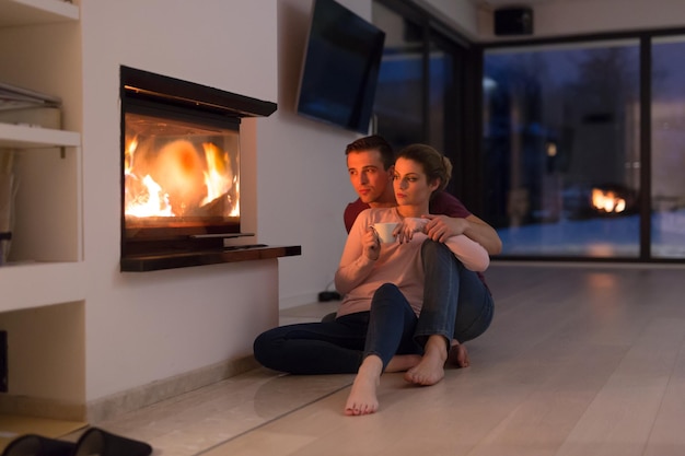 Junges romantisches Paar sitzt auf dem Boden vor dem Kamin, redet und trinkt Tee in der kalten Winternacht zu Hause