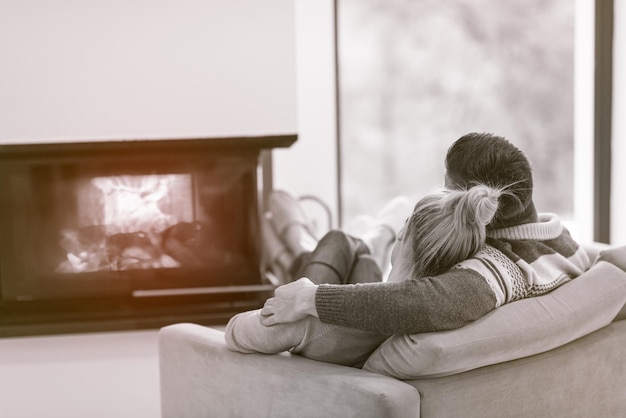 Junges romantisches Paar sitzt zu Hause auf dem Sofa vor dem Kamin, schaut sich an, redet und trinkt Kaffee an einem Herbsttag