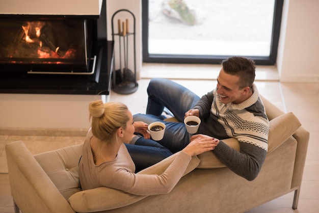 Junges romantisches Paar sitzt zu Hause auf dem Sofa vor dem Kamin, sieht sich an, redet und trinkt Kaffee am Herbsttag