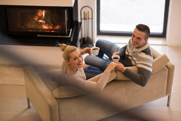 Junges romantisches Paar sitzt zu Hause auf dem Sofa vor dem Kamin, sieht sich an, redet und trinkt Kaffee am Herbsttag