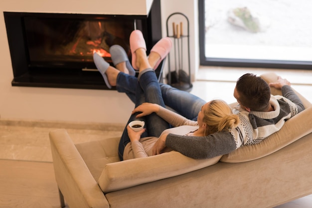 Junges romantisches Paar sitzt zu Hause auf dem Sofa vor dem Kamin, sieht sich an, redet und trinkt Kaffee am Herbsttag