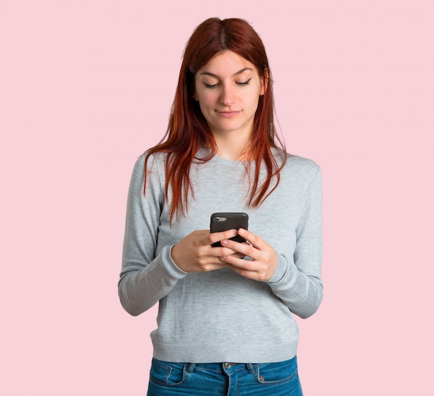 Junges Rothaarigemädchen, das eine Mitteilung oder eine E-Mail mit dem Mobile auf lokalisiertem rosa Hintergrund sendet