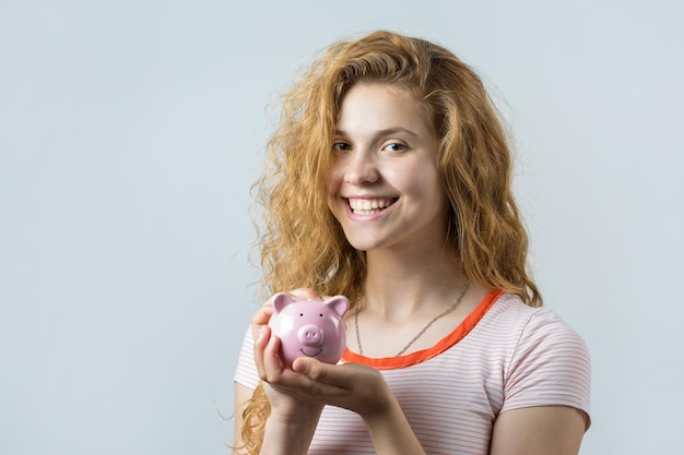 Junges rothaariges lockiges Mädchen über gelbem Hintergrund, das Sparschwein hält und freut sich gestikulierend Das Konzept der Zuverlässigkeit von Geldanlagen und Versicherungen