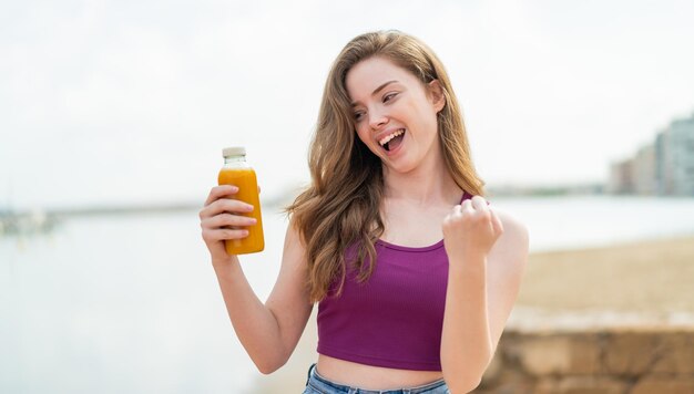 Junges rothaariges Mädchen hält draußen einen Orangensaft in der Hand und feiert einen Sieg