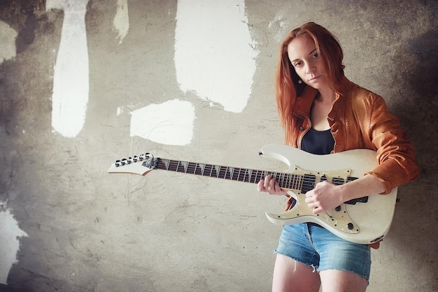 Junges rothaariges Mädchen mit E-Gitarre. Rockmusikermädchen in einer Lederjacke. Sie ist eine hübsche Sängerin und eine Darstellerin von Rockmusik.