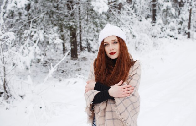 Junges rothaariges Mädchen mit langen roten Haaren auf einem Winterhintergrund. Rothaarige Frau in einem weißen Hut auf dem Hintergrund der Winternatur.