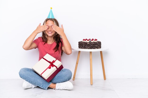 Junges russisches Mädchen, das einen Geburtstag feiert, sitzt auf dem Boden isoliert auf weißem Hintergrund und bedeckt die Augen mit den Händen