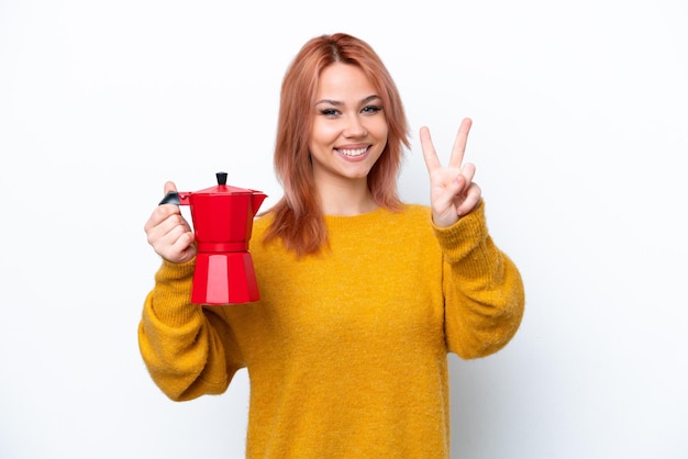 Junges russisches Mädchen, das Kaffeekanne isoliert auf weißem Hintergrund hält, lächelt und zeigt Victory-Zeichen