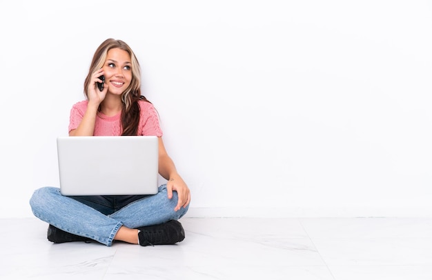 Junges russisches Mädchen mit einem Laptop, der auf dem Boden sitzt, isoliert auf weißem Hintergrund, der ein Gespräch mit dem Handy mit jemandem führt