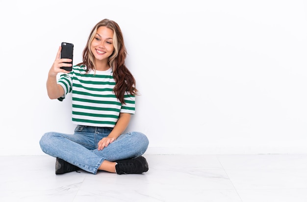 Junges russisches Mädchen sitzt auf dem Boden isoliert auf weißem Hintergrund und macht ein Selfie