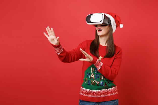 Junges Santa-Mädchen, das in Headset schaut, berührt so etwas wie Drücken Sie auf die Schaltfläche und zeigen Sie auf den schwebenden virtuellen Bildschirm, der auf rotem Hintergrund isoliert ist. Frohes neues Jahr 2019 Feier Urlaub Party Konzept.