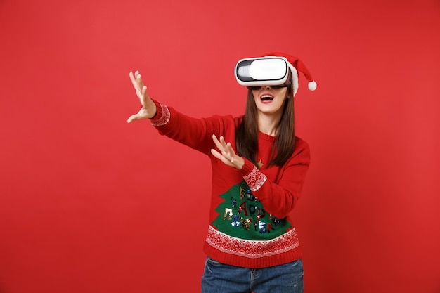 Junges Santa-Mädchen, das in Headset schaut, berührt so etwas wie Drücken Sie auf die Schaltfläche und zeigen Sie auf den schwebenden virtuellen Bildschirm, der auf rotem Hintergrund isoliert ist. Frohes neues Jahr 2019 Feier Urlaub Party Konzept.