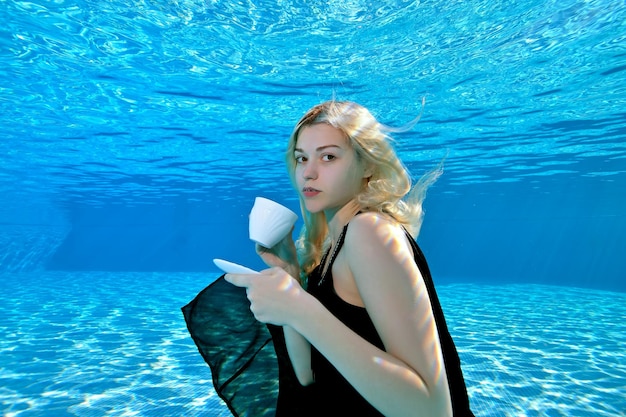 Junges schönes blondes Mädchen, das unter Wasser im Pool mit einer weißen Kaffeetasse in ihren Händen posiert