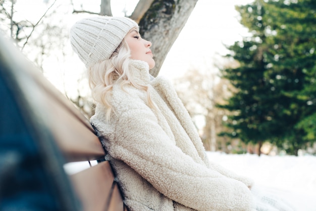 Junges schönes blondes Mädchen sitzt auf einer Bank im Park im Winter