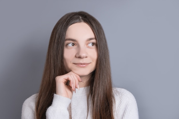 Junges schönes Frauenporträt mit dunklem Haar, das weißen Pullover trägt, schaut weg mit einem leichten Lächeln .Studio schoss auf grauem Hintergrund.