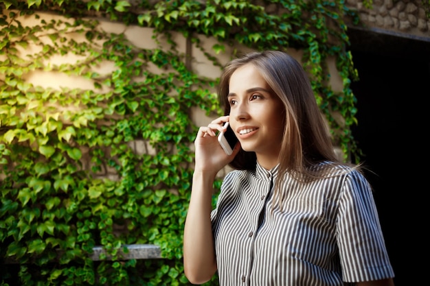 Junges schönes fröhliches Mädchen, das durch die Stadt geht und am Telefon spricht