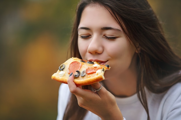 Junges schönes fröhliches Mädchen, das Pizza isst, während es den Geschmack in der Natur genießt