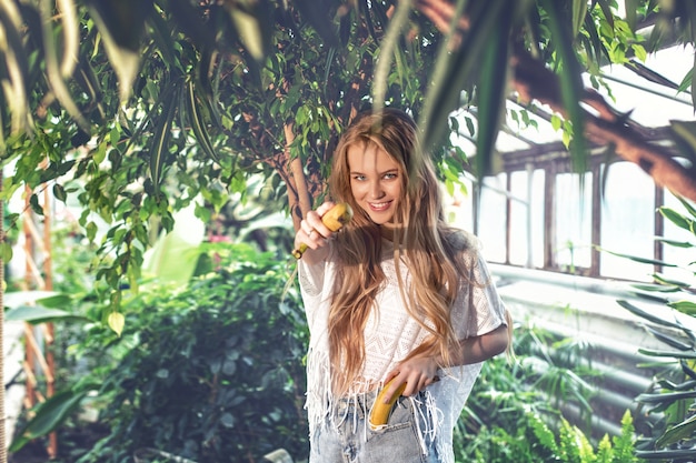 Junges schönes glückliches blondes Mädchenmodell mit Bananen auf tropischem Pflanzenhintergrund