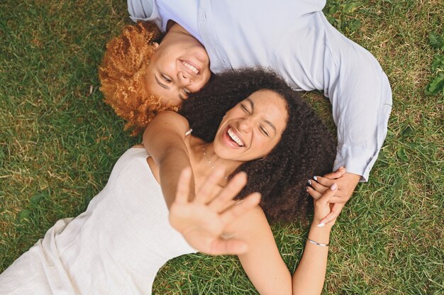 Junges schönes glückliches lesbisches afroamerikanisches Paar, das auf Gras liegt und lachend die Hände ausstreckt