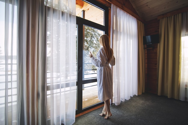 Junges schönes glückliches Mädchen in einem Morgenmantel steht an einem großen Fenster mit einem Glas