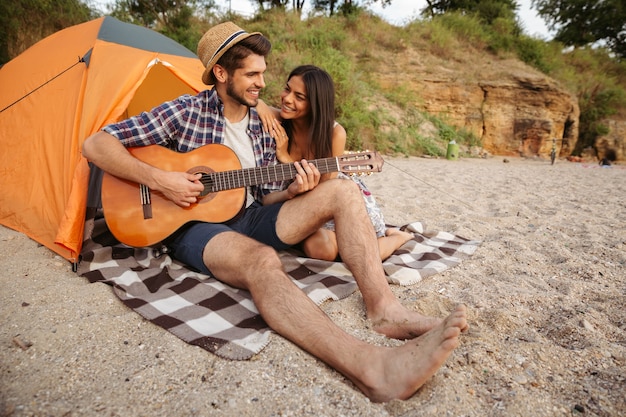 Junges schönes glückliches Paar, das Gitarre spielt, während es am Strand sitzt