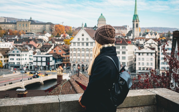 Junges schönes hübsches blondes Touristenmädchen in warmem Hut und Mantel mit Rucksack, das im kalten Herbst in der europäischen Stadt spazieren geht und ihre Reise in Zürich, Schweiz, genießt