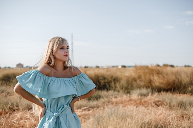 Junges schönes kaukasisches blondes Mädchen im hellblauen Kleid steht auf dem Feld mit dem sonnenversengten Gras