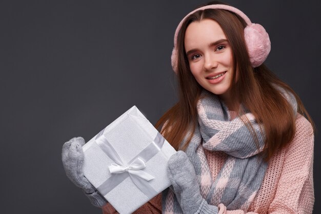 Junges schönes lächelndes Mädchen beim Stricken des Pullovers und der rosa flaumigen Ohrenschützer, die Geschenkbox halten