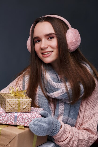 Junges schönes lächelndes Mädchen beim Stricken des Pullovers und der rosa flaumigen Ohrenschützer, die Geschenkboxen halten