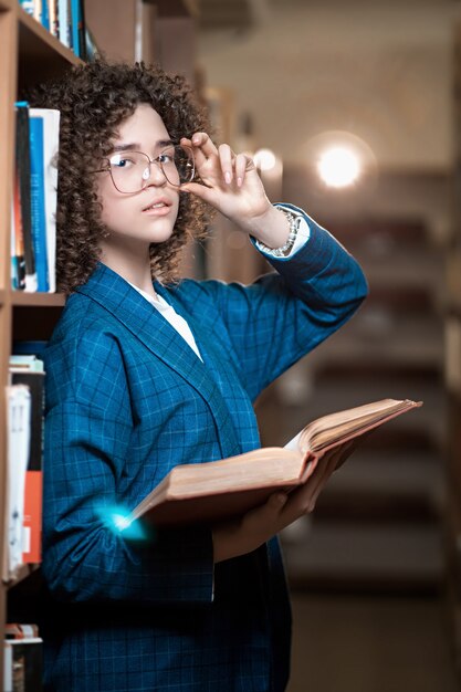 Junges schönes lockiges Mädchen in den Gläsern und im blauen Anzug steht in der Bibliothek